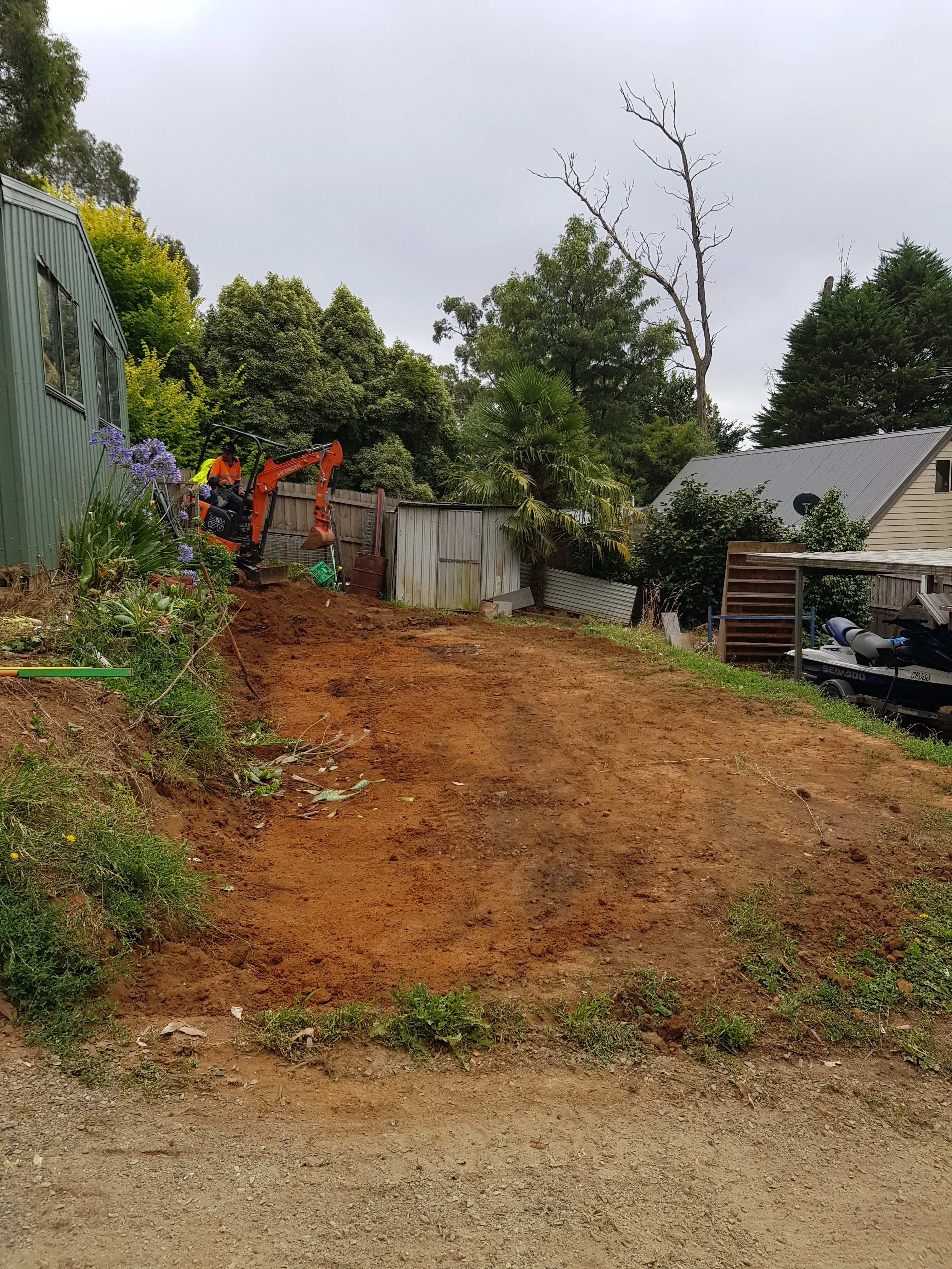 Freshly excavated red clay embankment in a backyard with a green weatherboard house and trees.