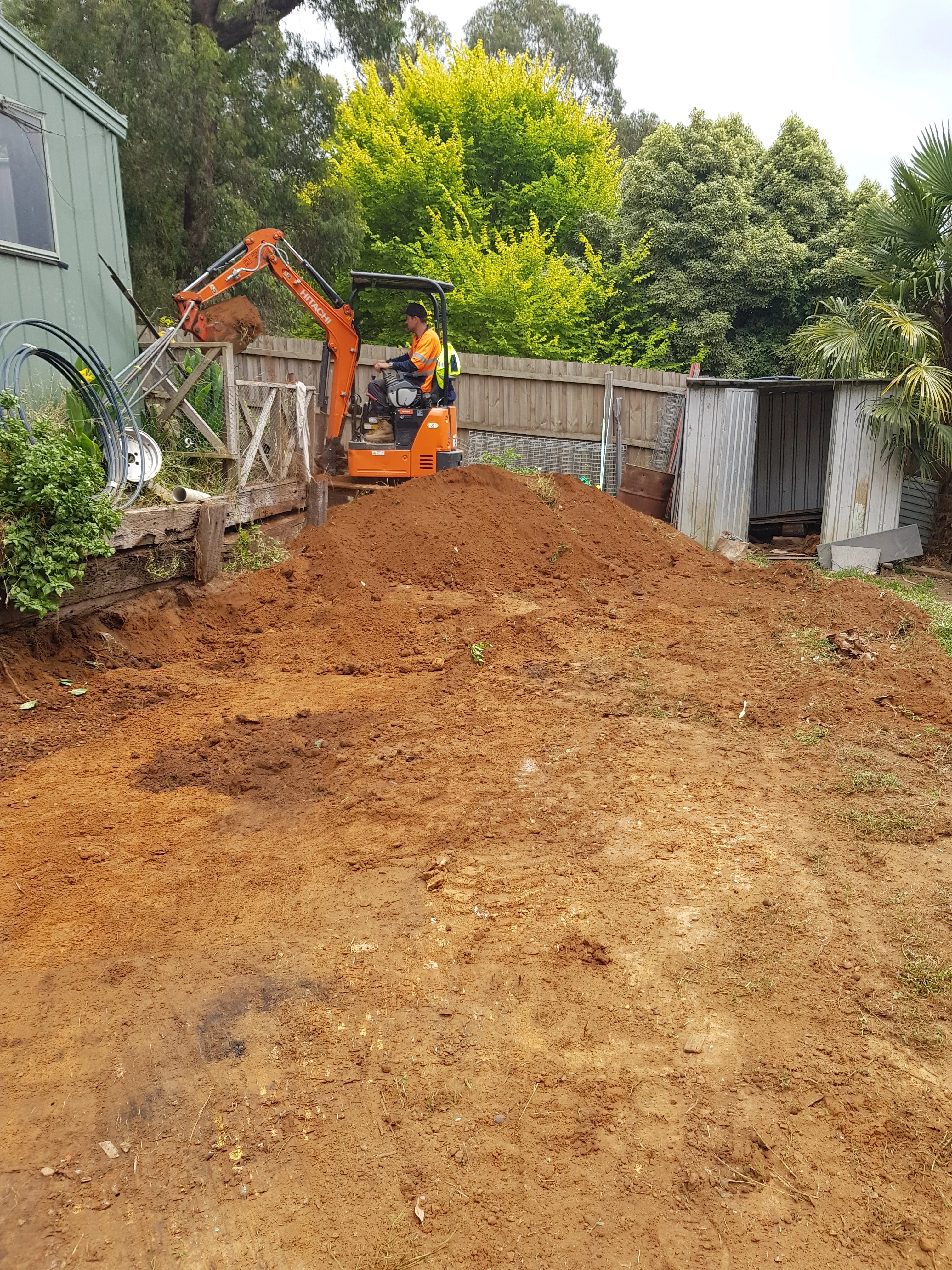 Cleared red clay slope after excavation, with an orange mini excavator at the top beside a shed.