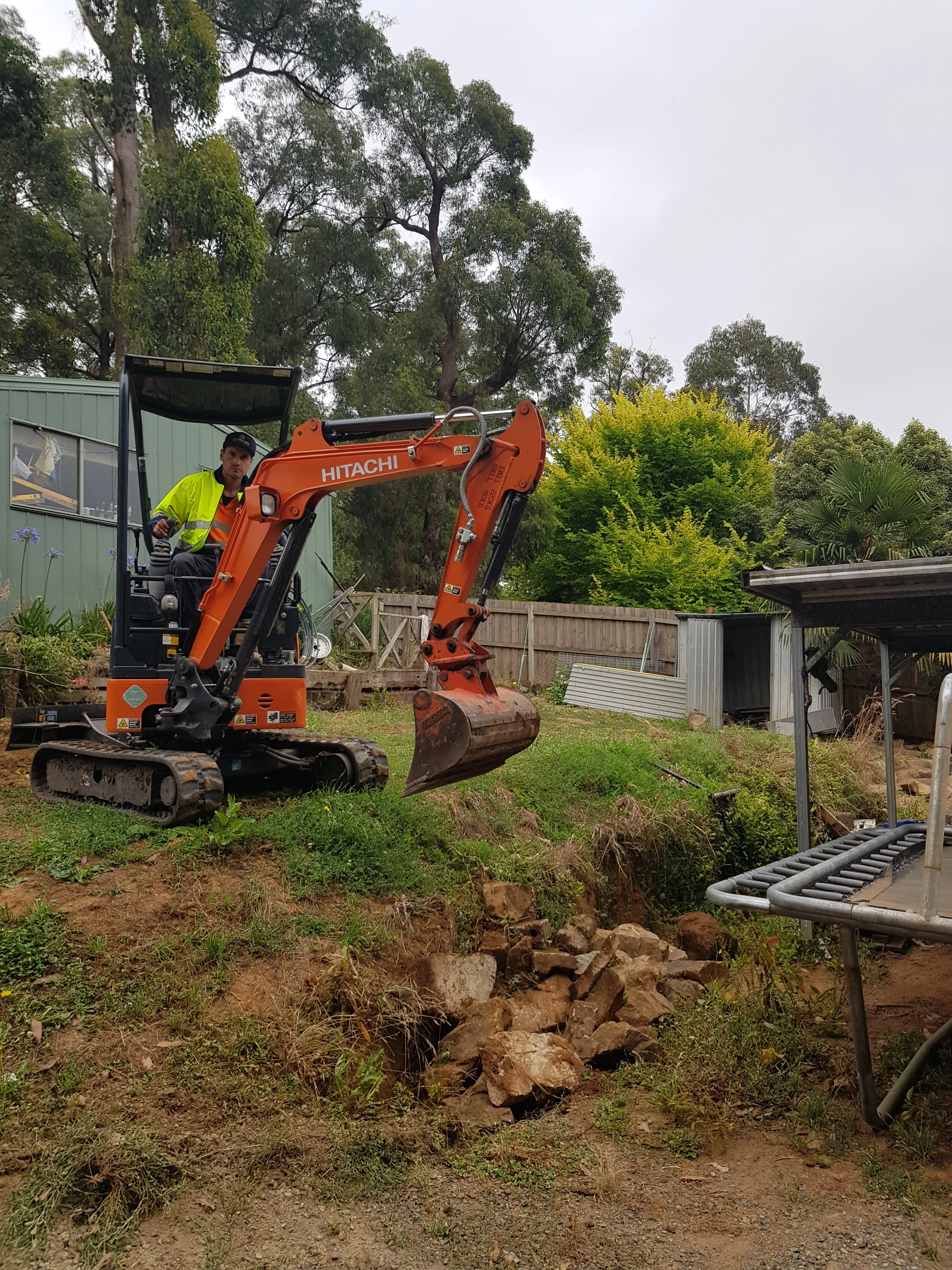 Orange mini excavator digging out a grassy backyard slope next to a timber fence.