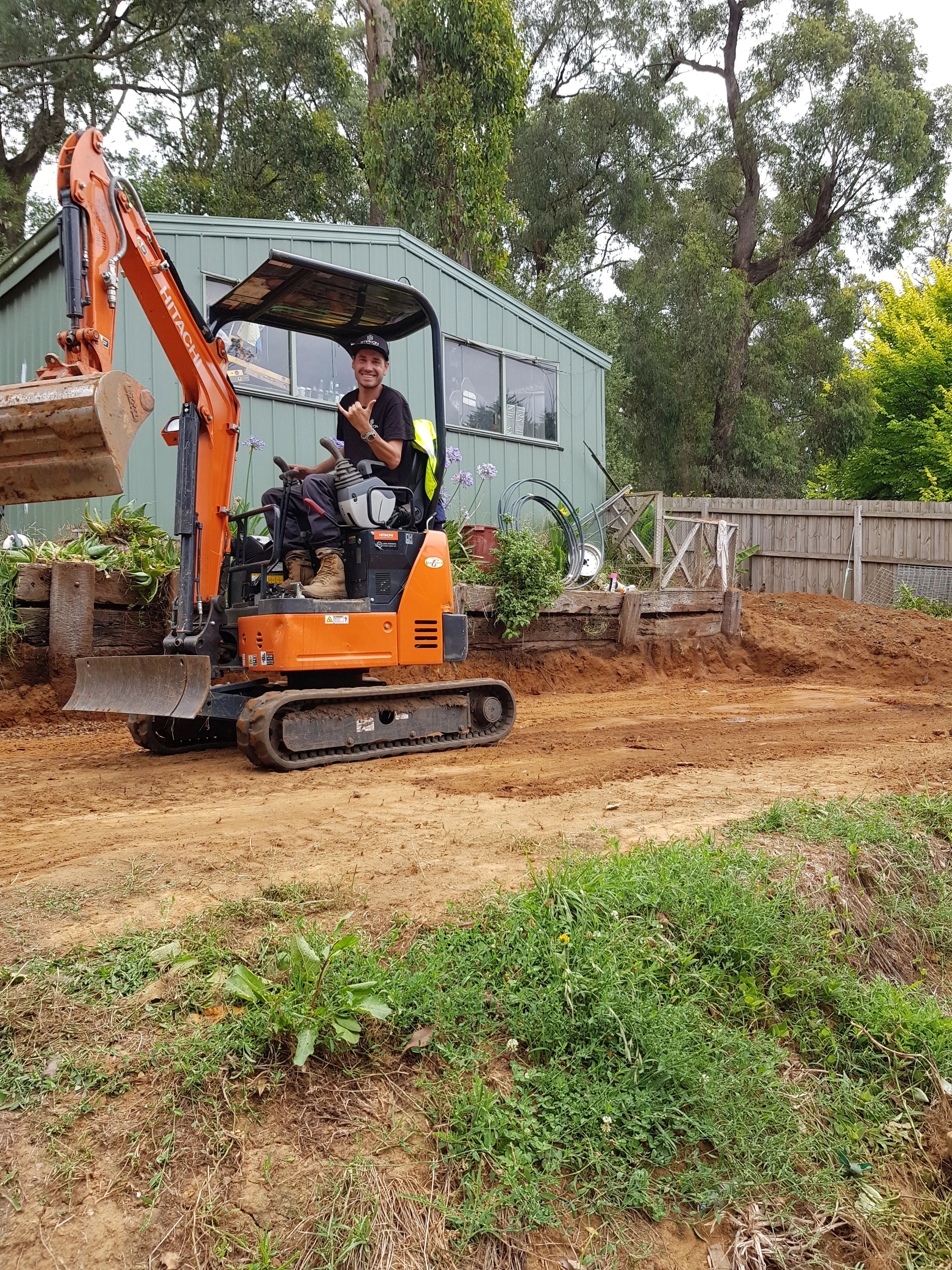 Operator seated in an orange mini excavator on a levelled clay pad next to a green weatherboard house.