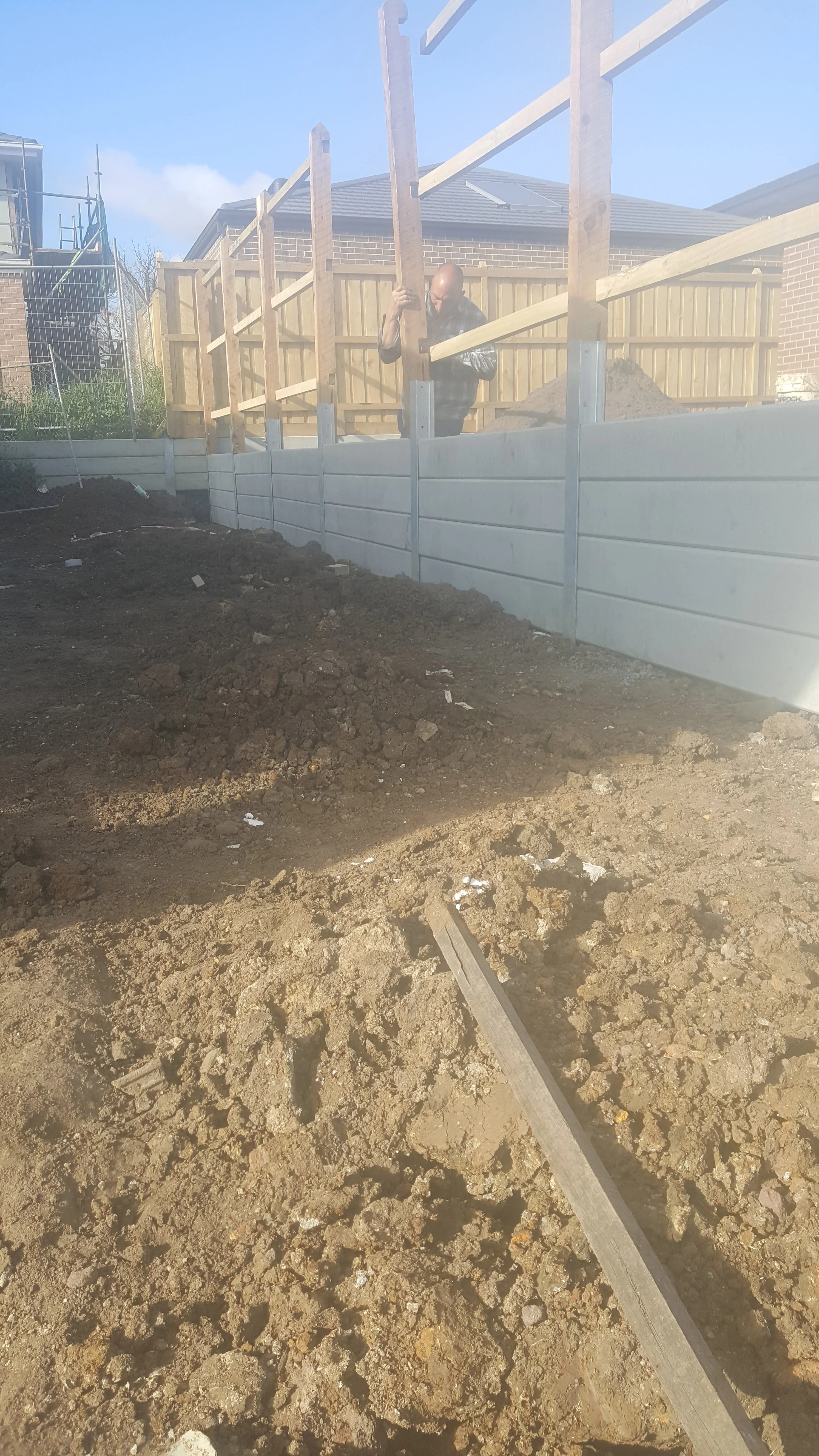 Grey concrete sleeper retaining wall along a boundary below a timber fence frame, with levelled soil in front.