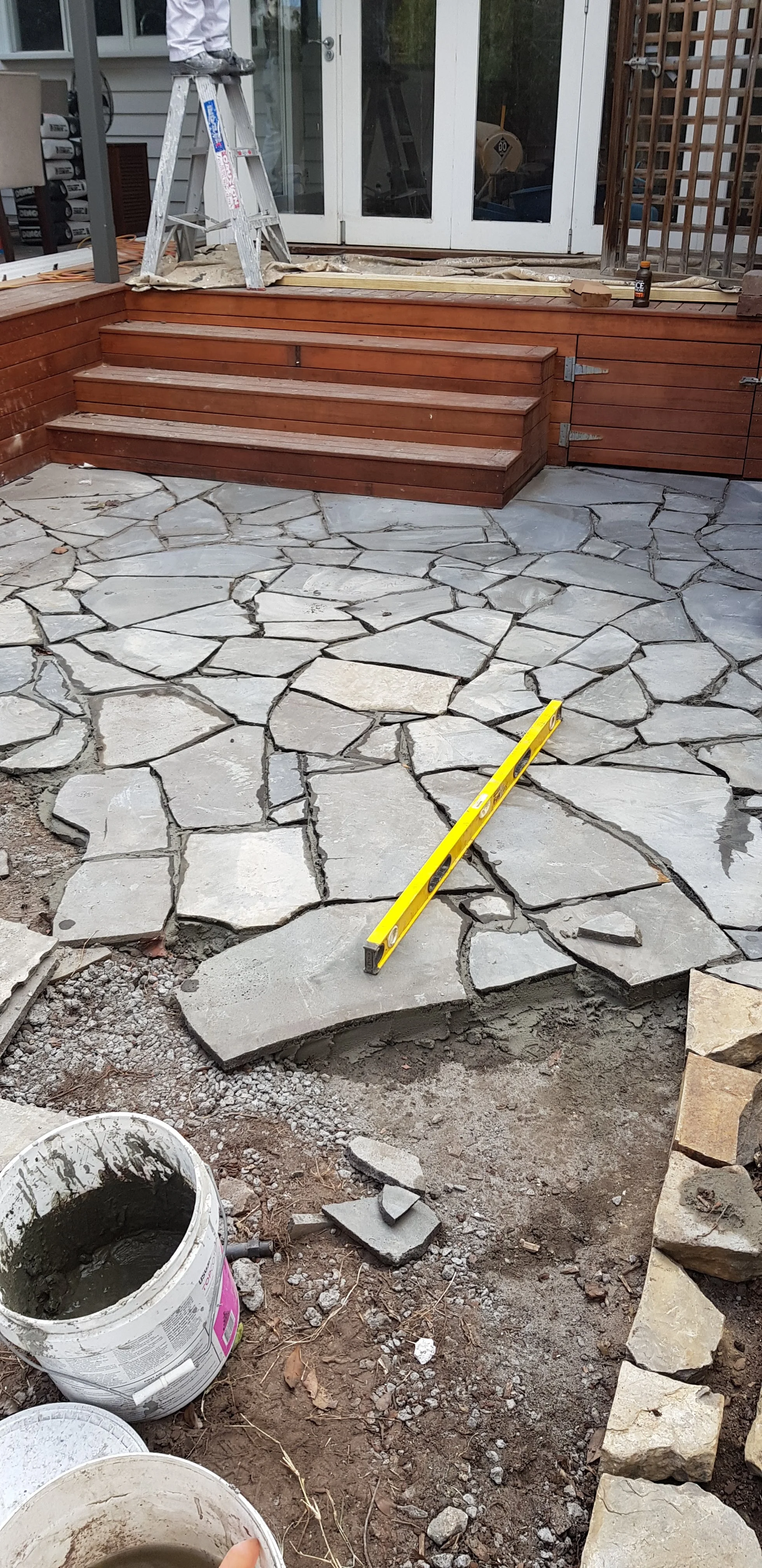 Bluestone crazy paving being laid on mortar with a yellow level, a mortar bucket and red timber steps.