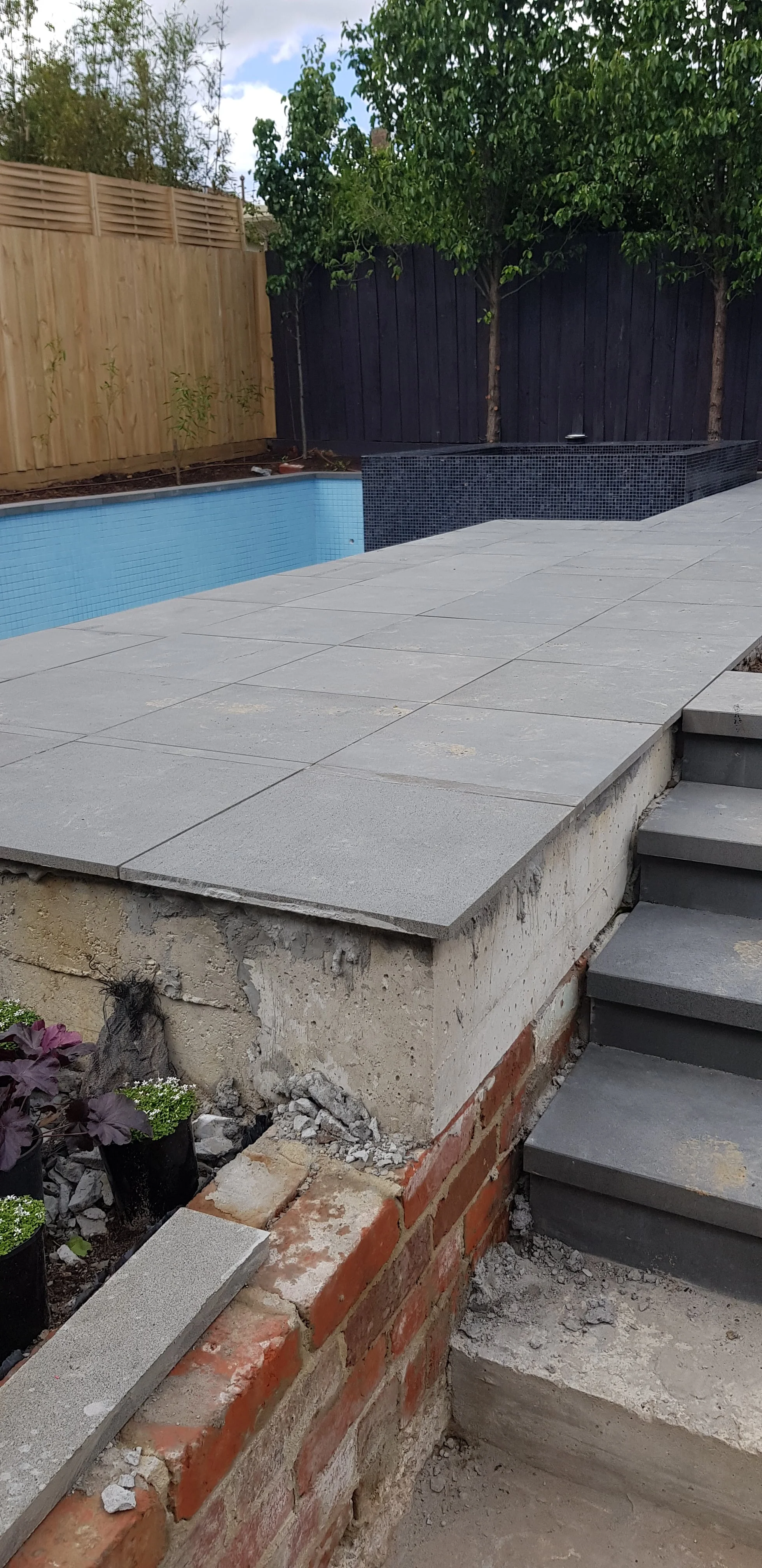 A grey paved landing and steps built over a brick base beside a pool.
