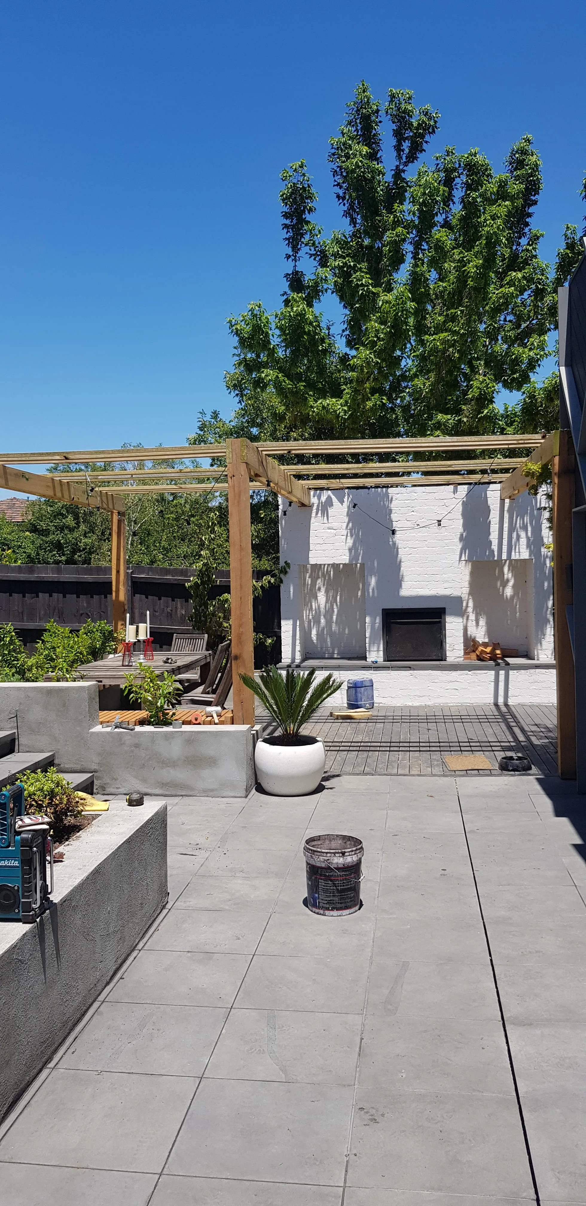 Paved courtyard of large grey tiles leading to a timber pergola, a white outdoor fireplace and raised garden beds under a blue sky.