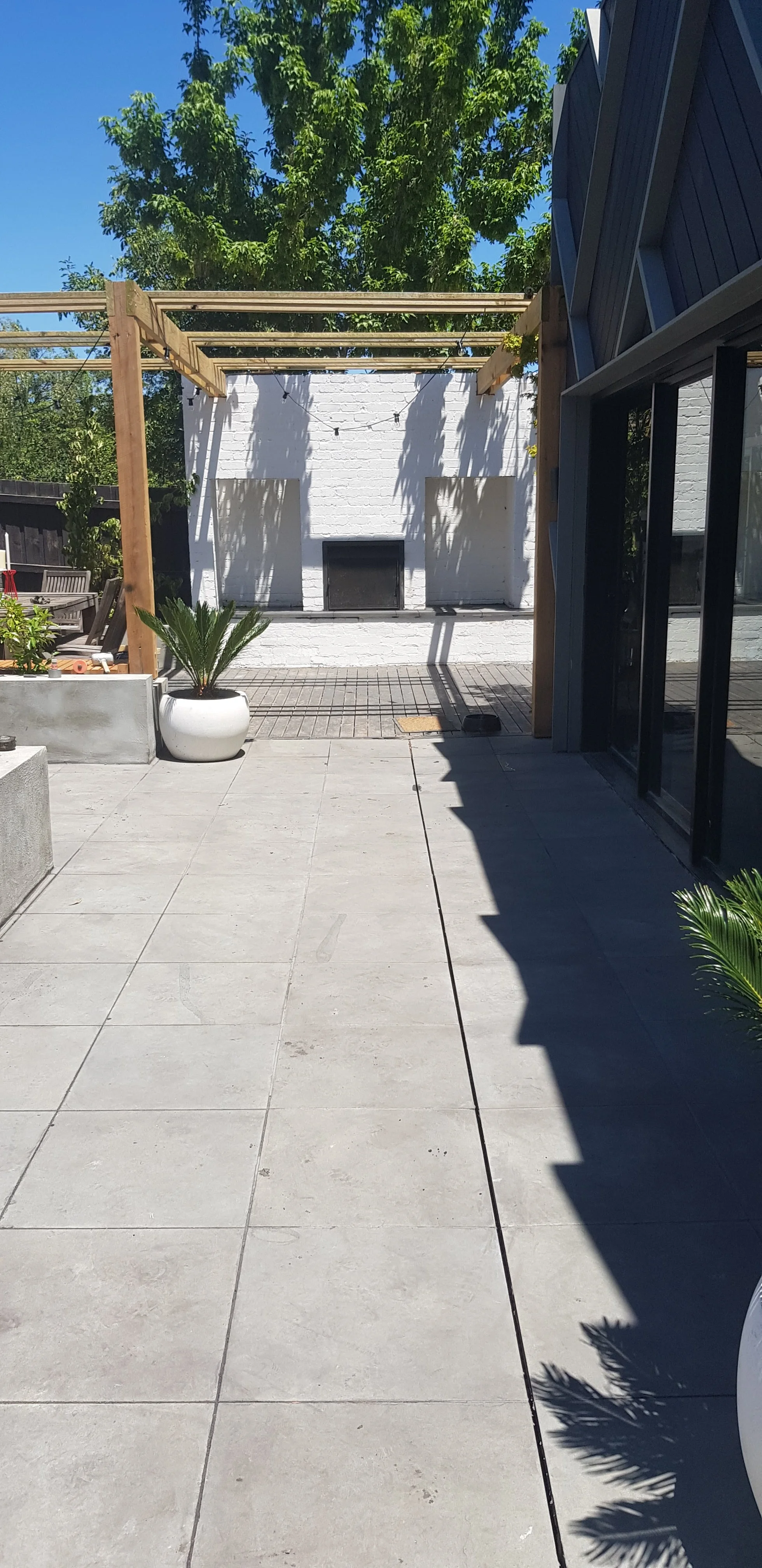 A wide grey paved side path leading to a timber pergola and outdoor fireplace.