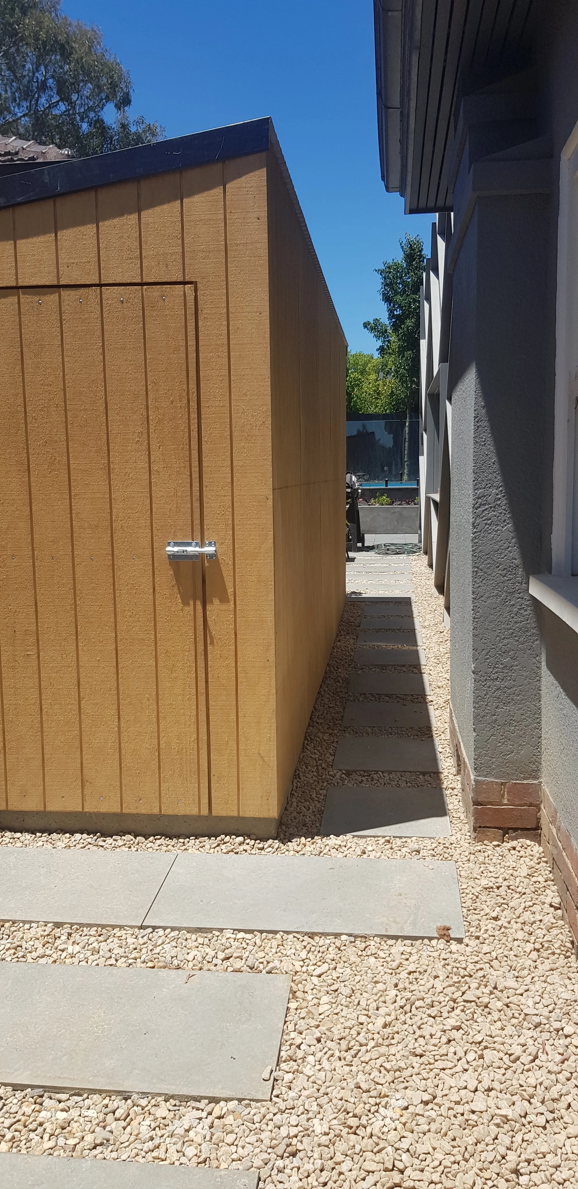 Pavers set in gravel leading to a timber-clad garden shed beside a dark house wall.