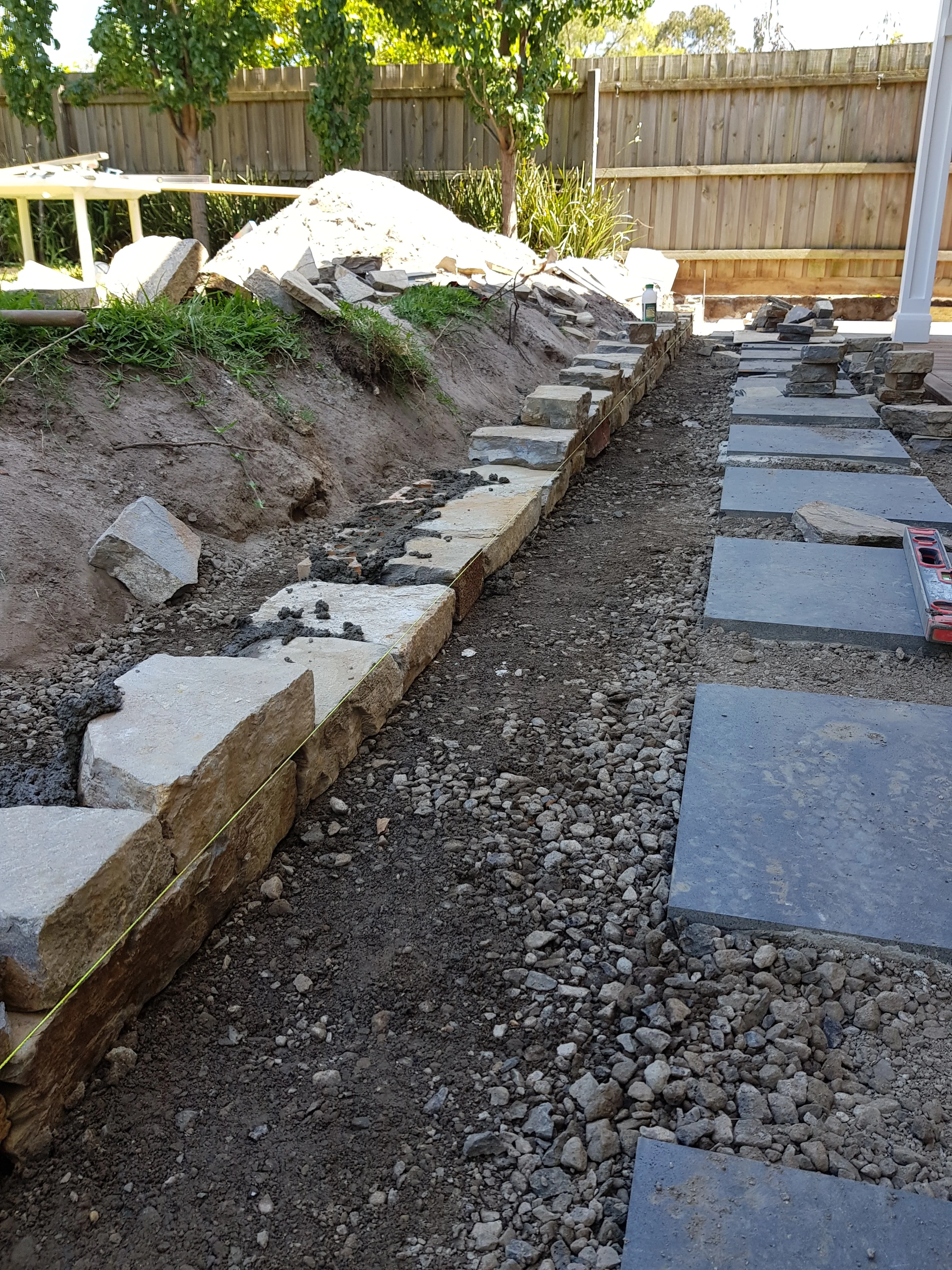 Dark bluestone stepping stones laid on a crushed-rock base between a stone-edged garden bed and timber decking.