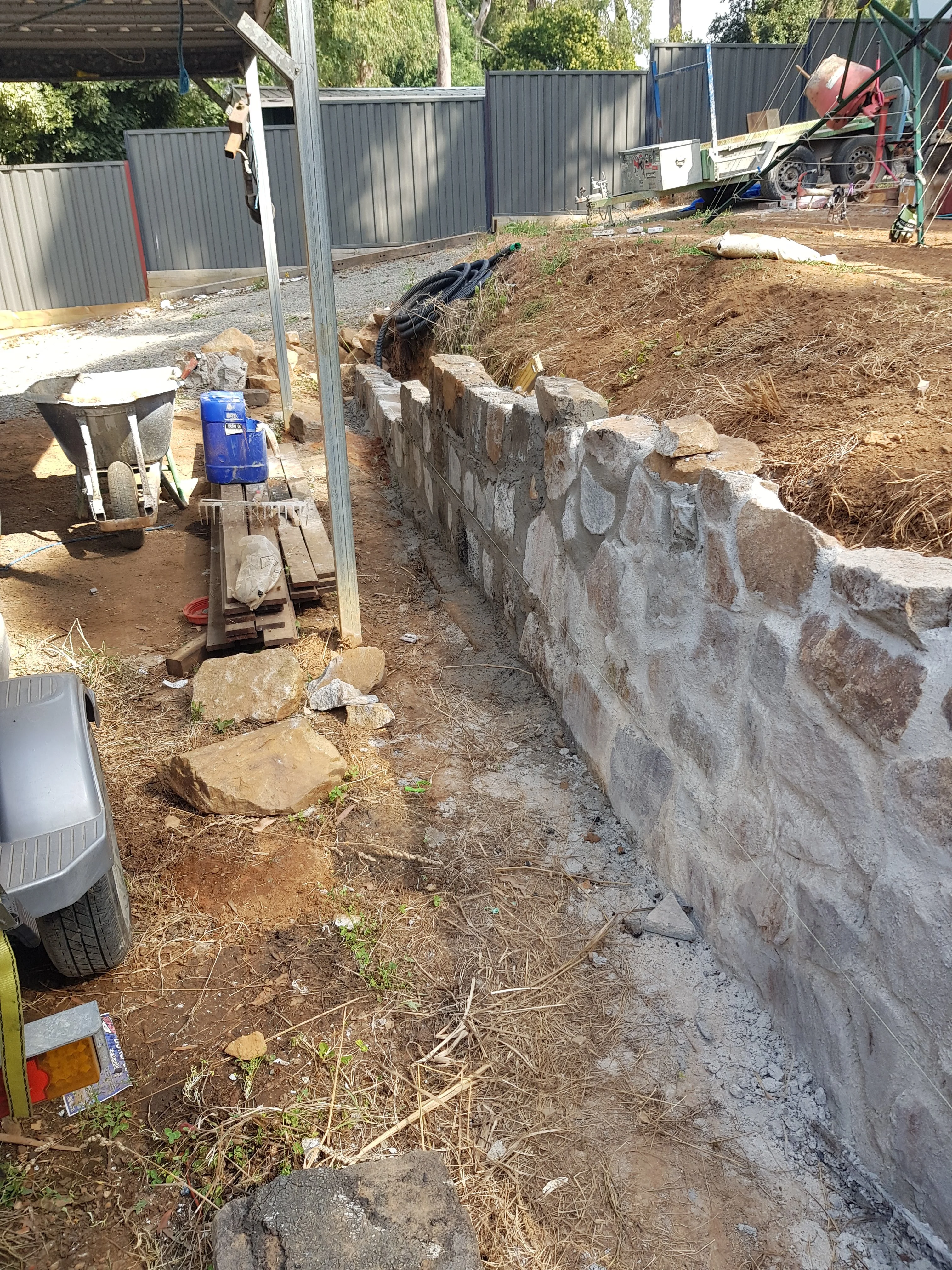 Stacked stone retaining wall along a narrow side path under construction, with tools and materials.
