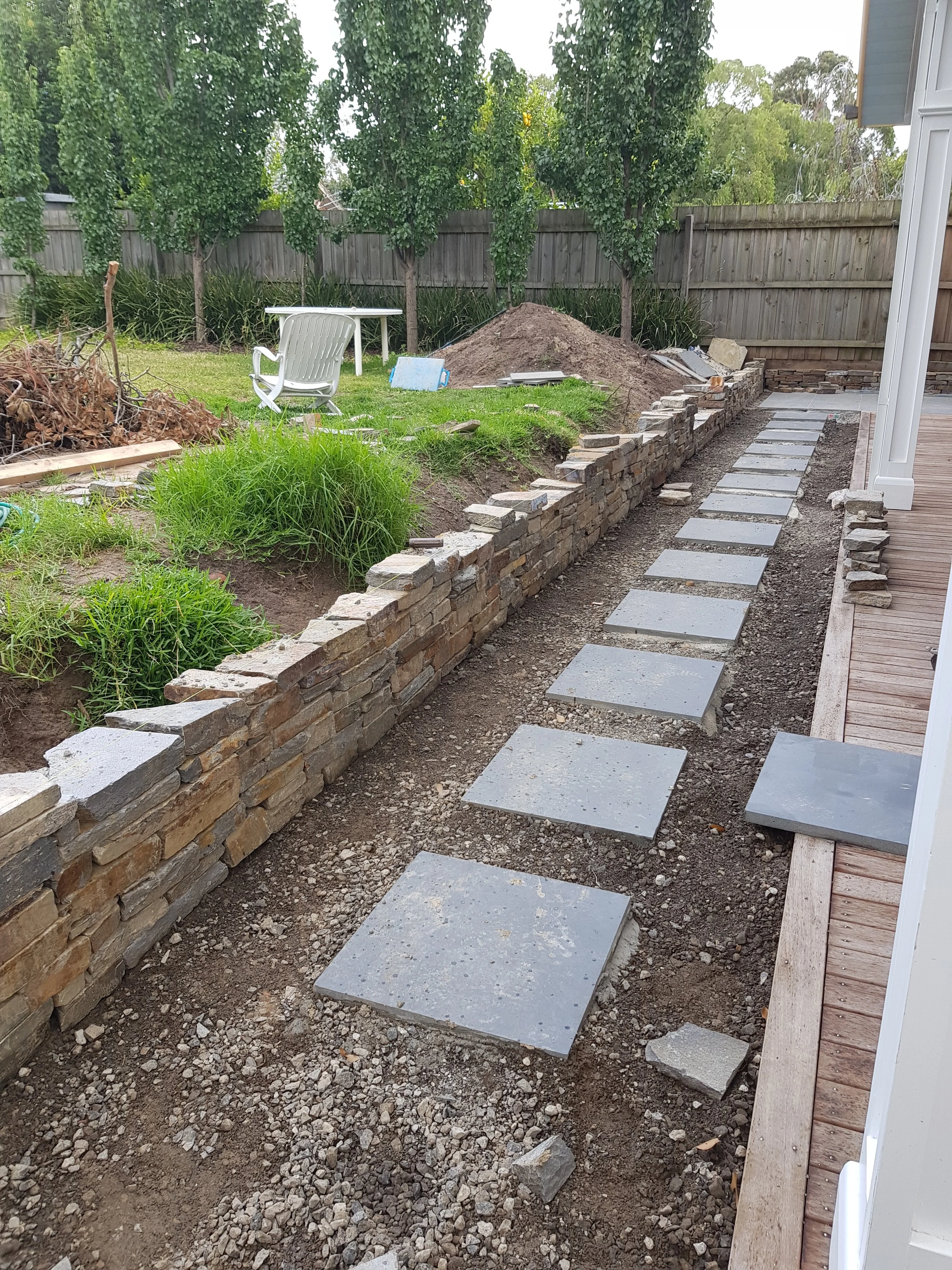Bluestone stepping stone path laid between a low stacked stone wall and timber decking, with the garden behind.