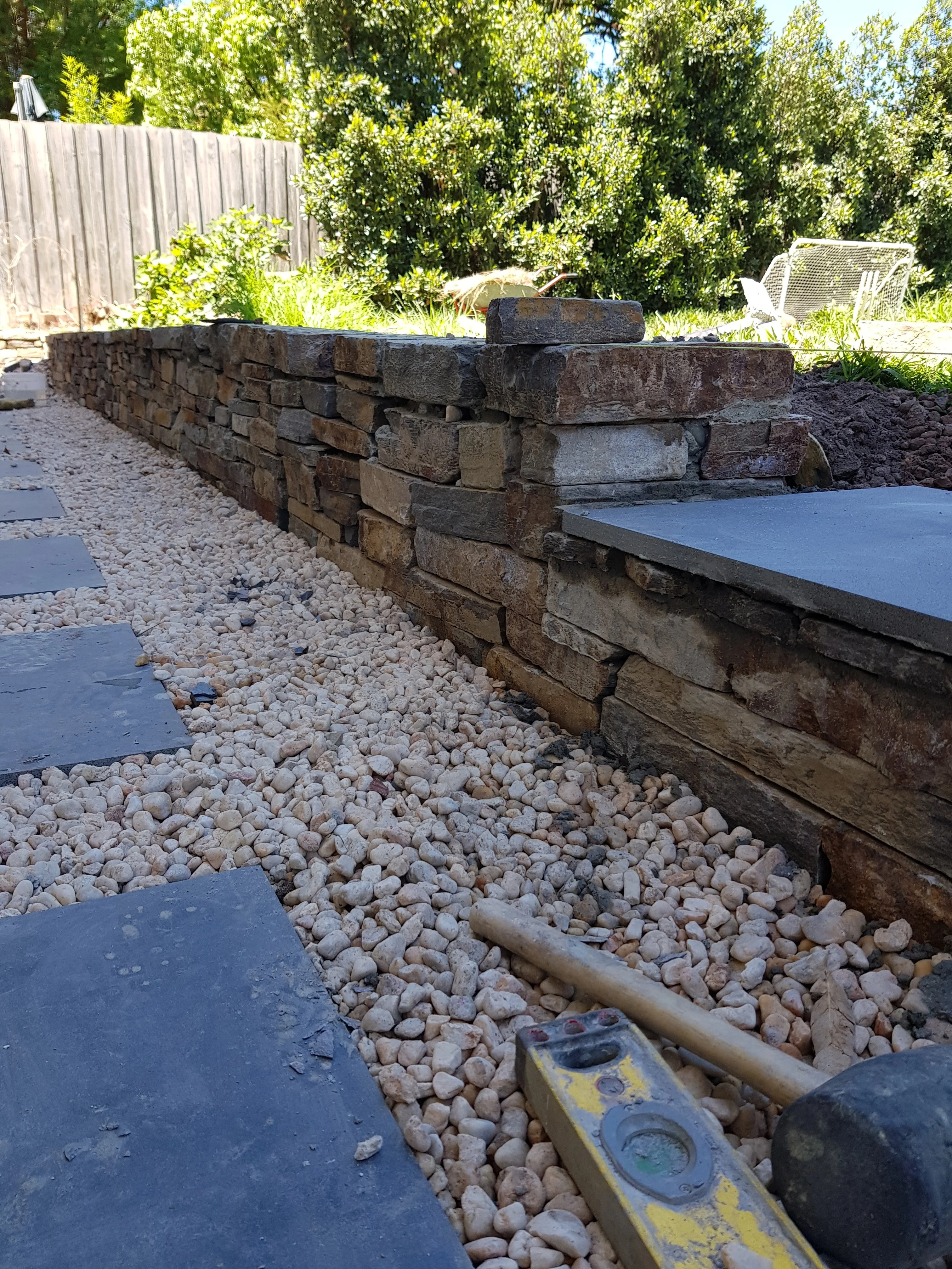 Dry stacked stone wall with a bluestone capping slab beside a pebble path, with a level and mallet on the path.