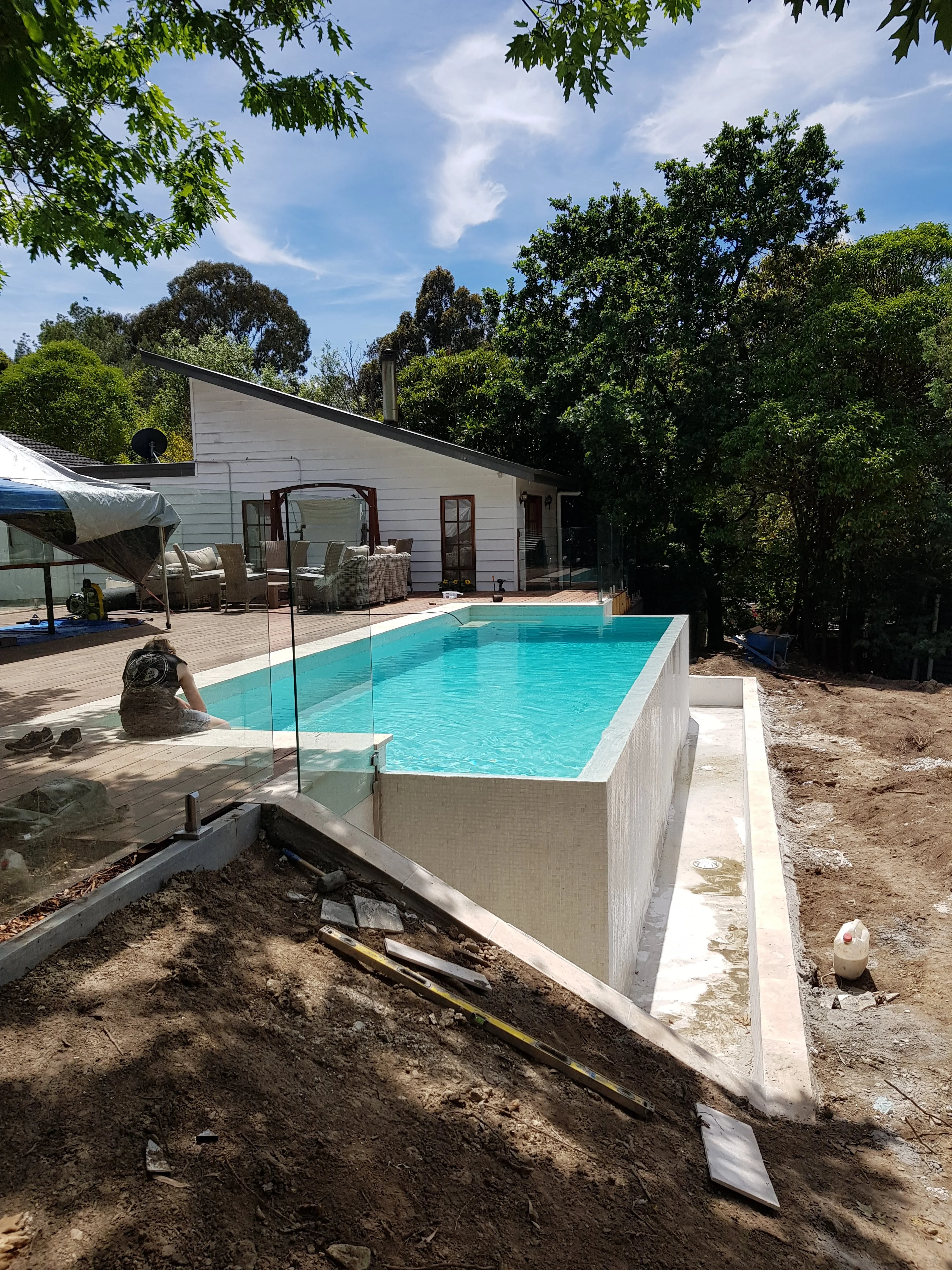 Raised concrete pool filled with clear blue water, with frameless glass fencing beside a white weatherboard house.