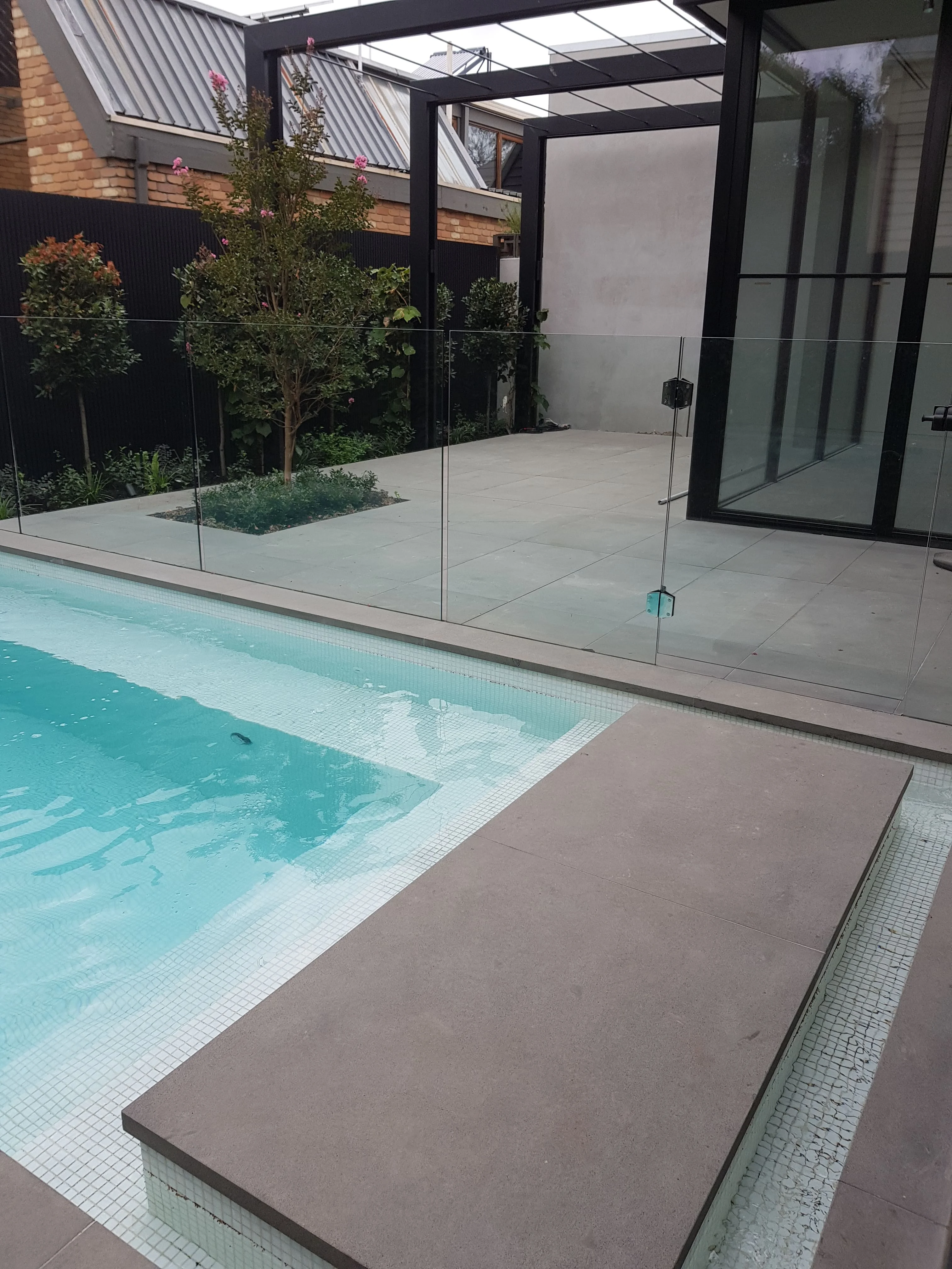Pool corner with wide dark coping and pale paving leading to a modern home with a black pergola.