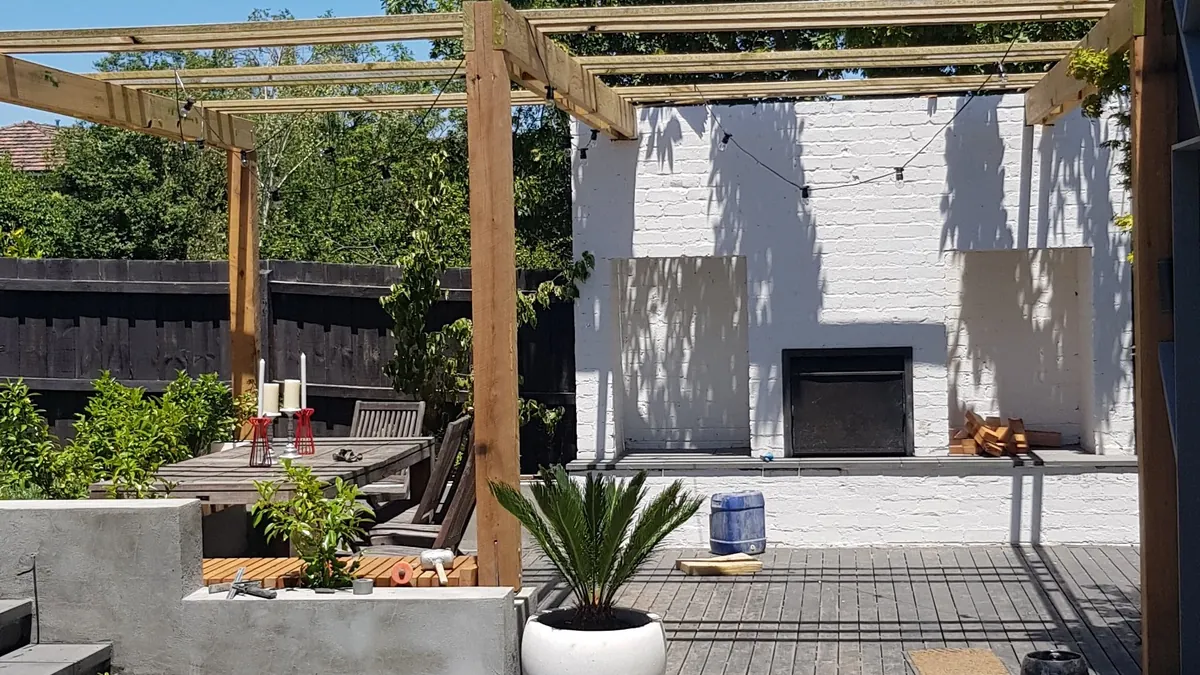 Large-format paved courtyard with raised planter, potted plants and a timber pergola under blue sky